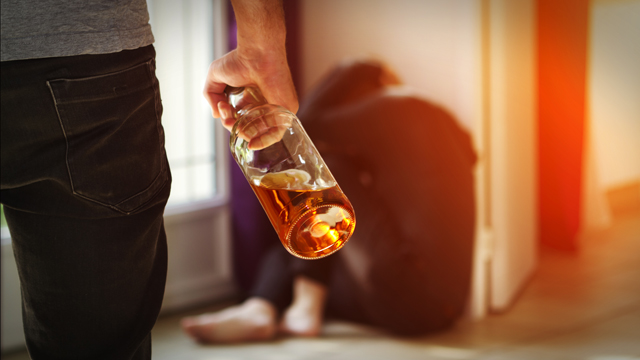 Man with bottle of alcohol and woman cowering against a wall