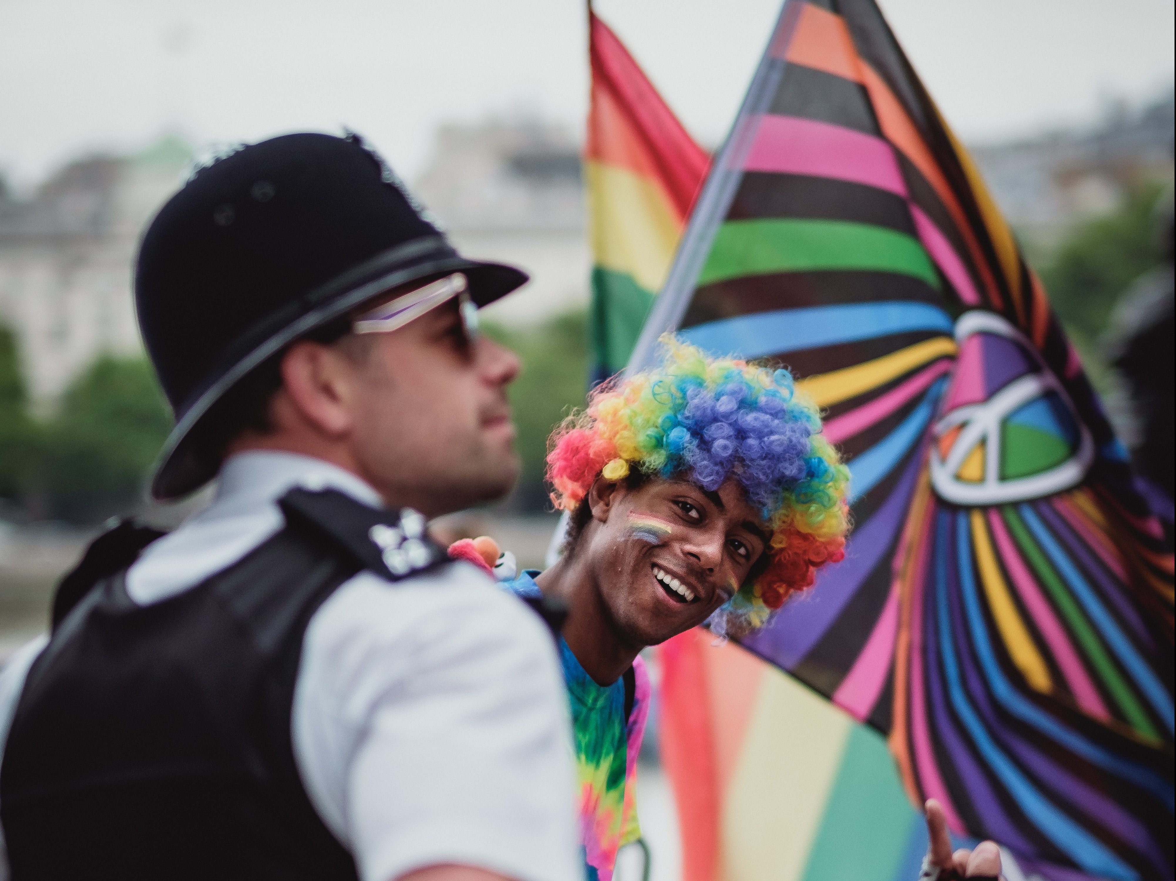 young man in a rainbow wig smiling at the camera, looking past a police officer in sunglasses