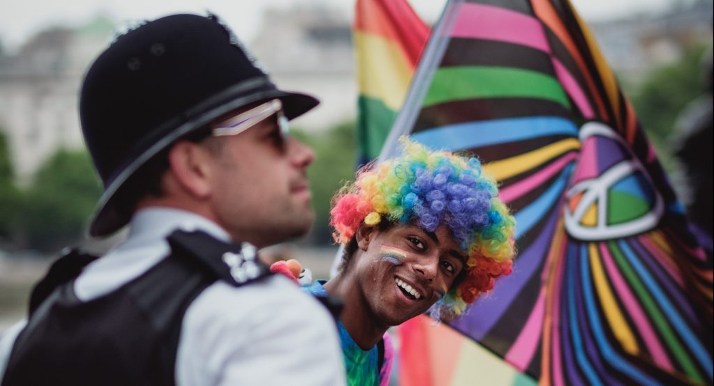 Police officer looking right in front of young black man in a rainbow wig, smiling and looking at the viewer.