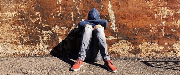 Teenager sat against a wall with their knees drawn up, arms folded, and their head resting on their arms.
