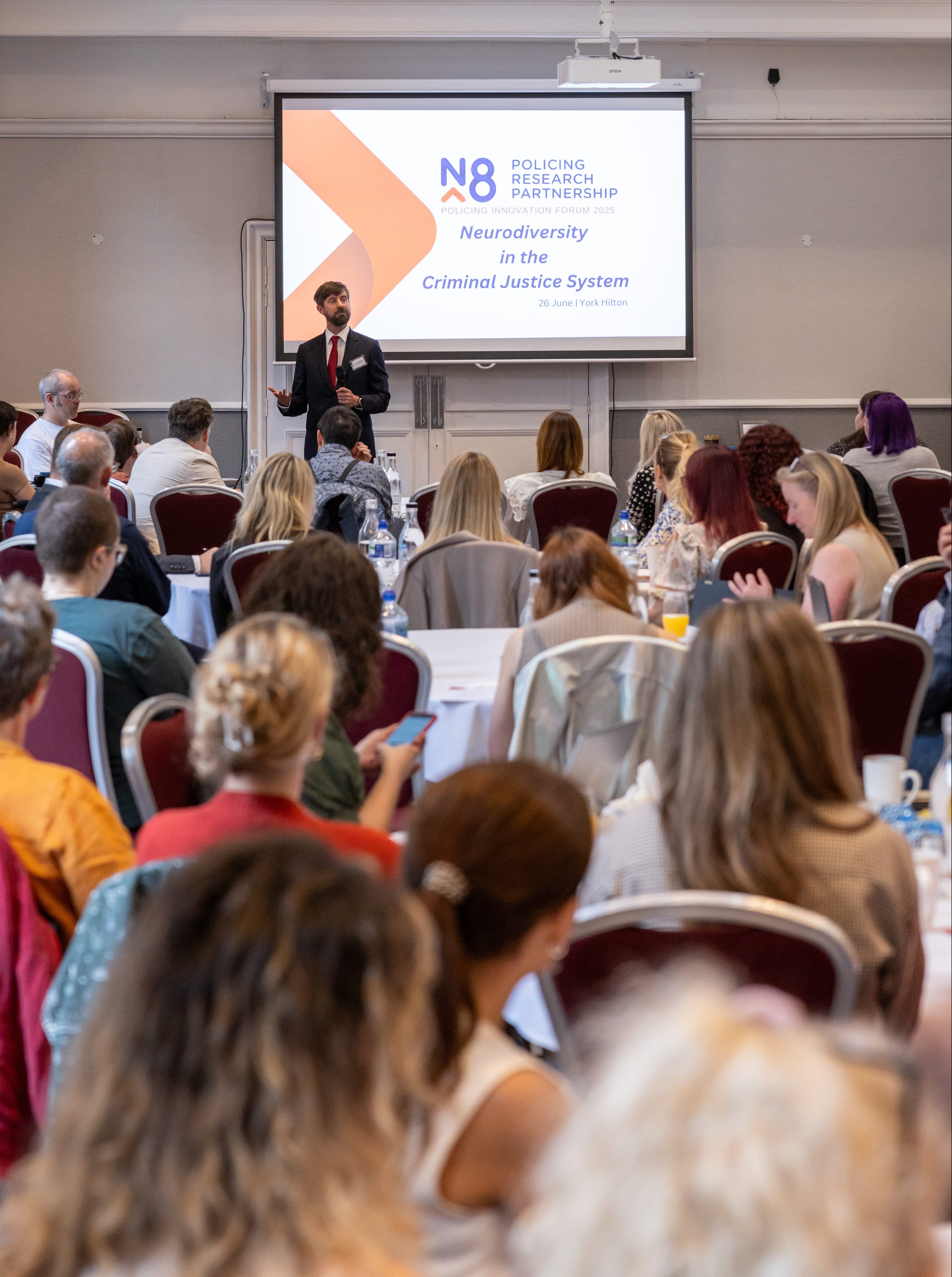 Man in a conference room addresses an audience sat at round tables
