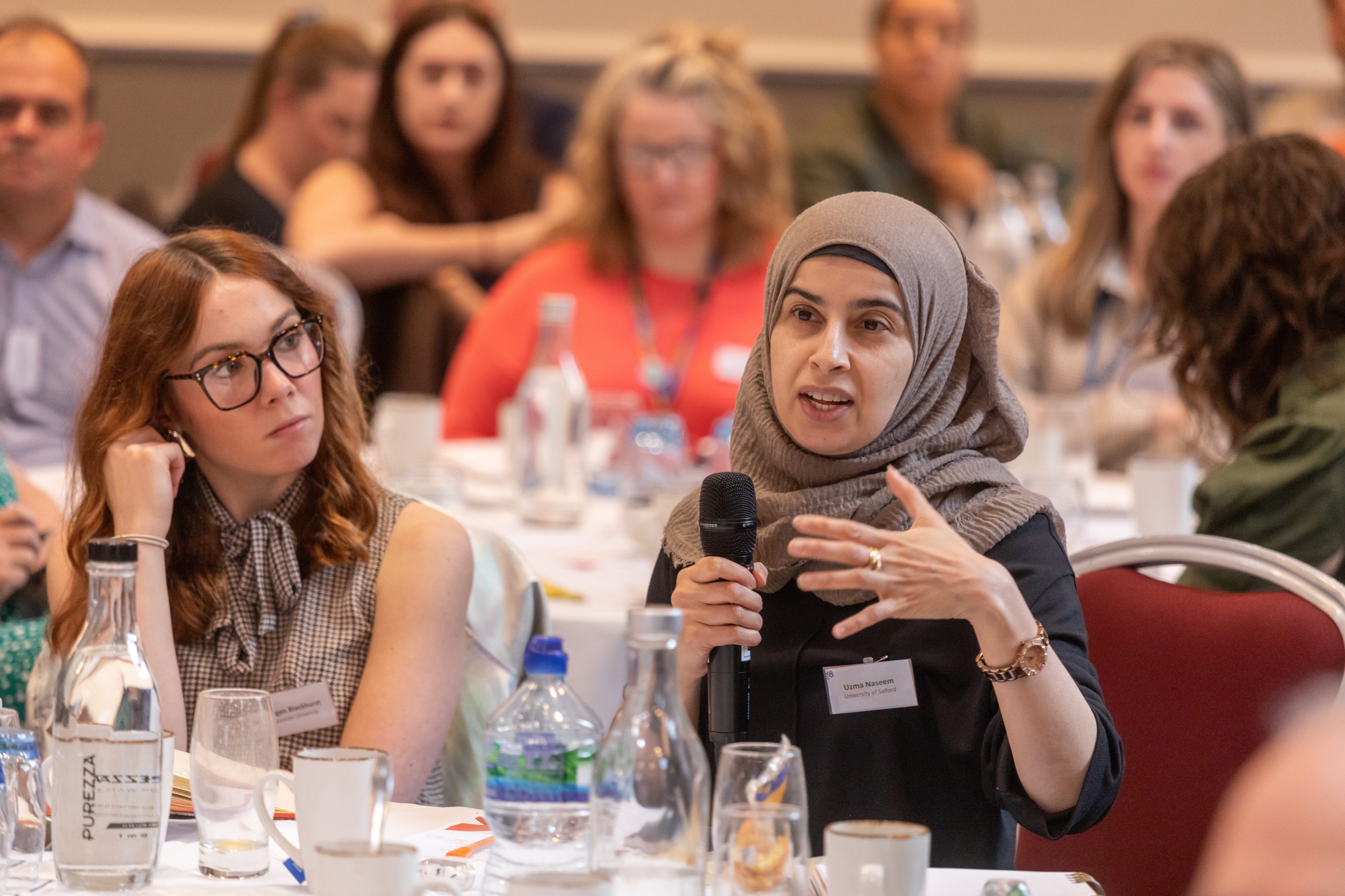 Woman speaking into a microphone sat at a table with other people, with further people sat at tables in the background